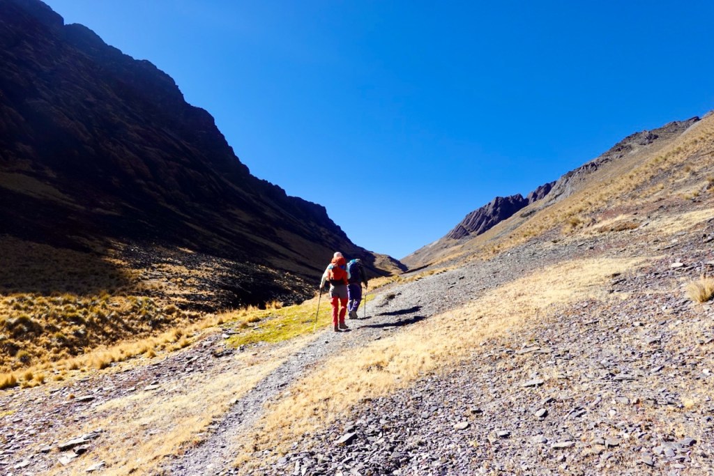 Two hikers walking on a rocky path surrounded by tall mountains under a clear blue sky.