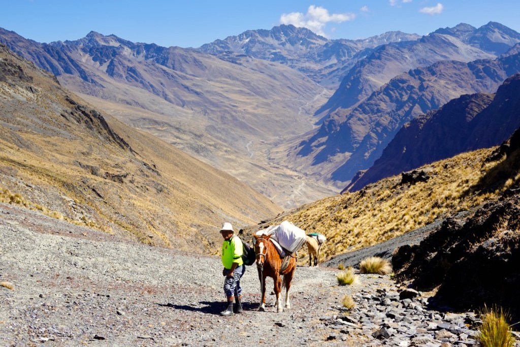 A person walking alongside a horse carrying a pack, with rugged mountains and a valley in the background.