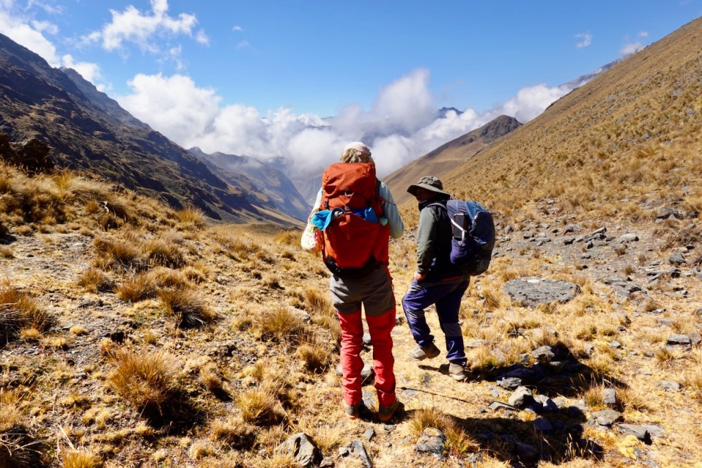 Two hikers with backpacks walking through a mountainous landscape with grassy terrain and clouds in the sky.