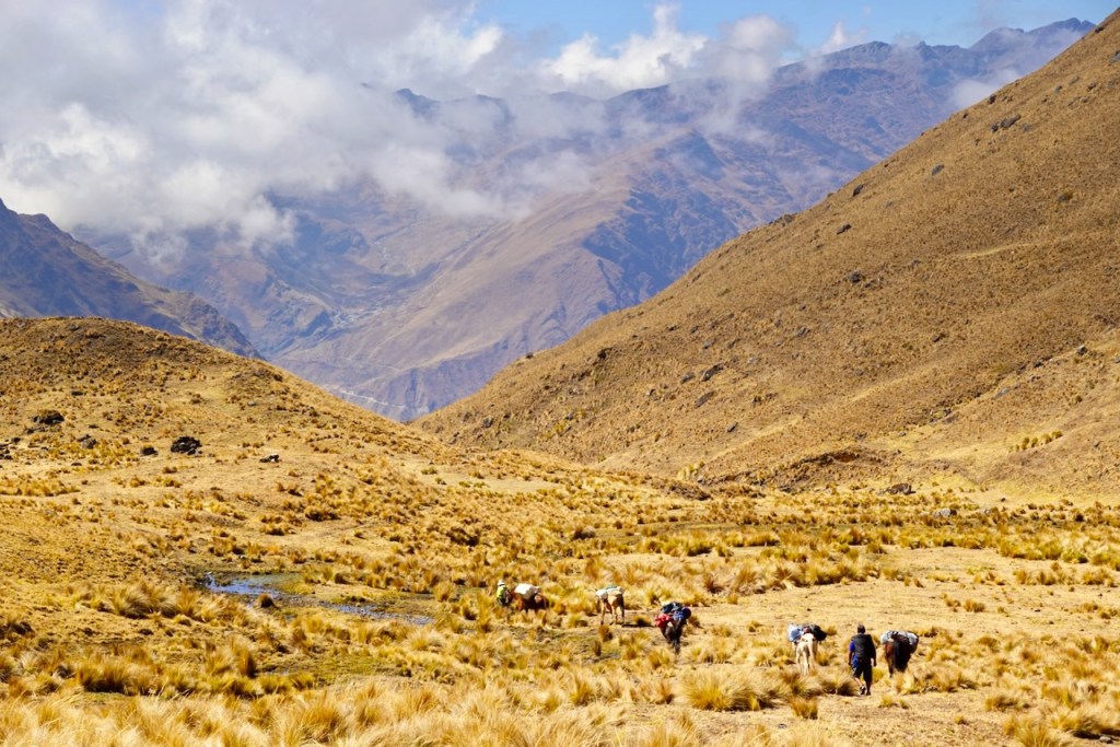 A group of people hiking through a grassy valley surrounded by mountains under a partly cloudy sky.