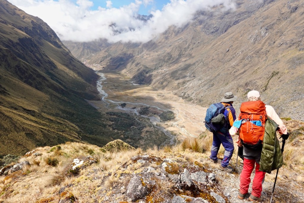 Two hikers standing on a mountain ridge overlooking a valley with a river and lush terrain under a partly cloudy sky.