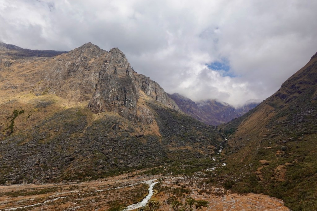 A panoramic view of a mountainous landscape featuring rugged terrain, steep cliffs, and a winding river flowing through a valley under a partly cloudy sky.