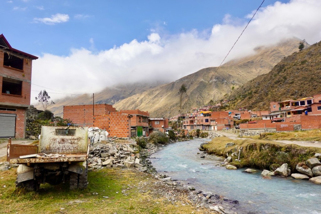 A scenic view of a small village in the mountains, featuring adobe houses, a meandering river, and steep hills under a cloudy sky.