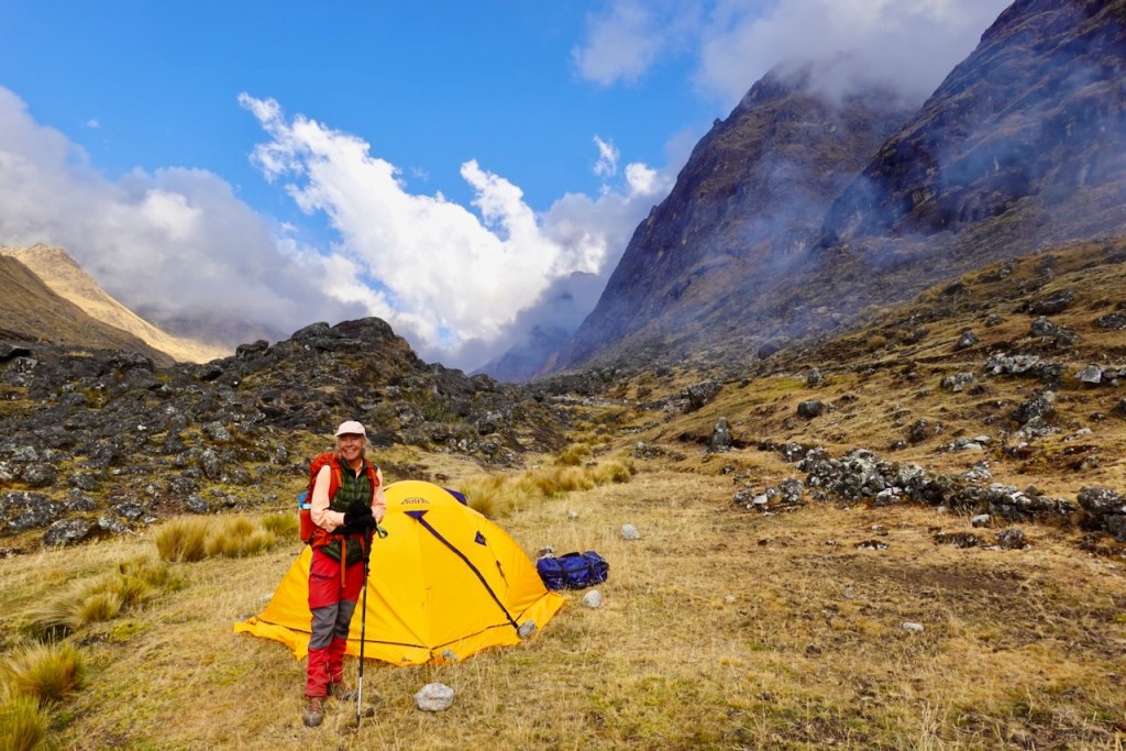 A person stands next to a yellow tent in a mountainous landscape, with rocky terrain and clouds in the background.