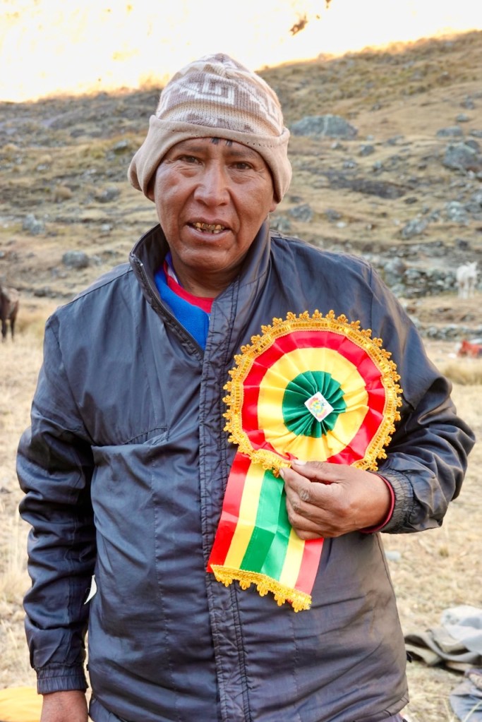 Man in a dark jacket holding a colorful rosette ribbon outdoors, with a mountainous background.