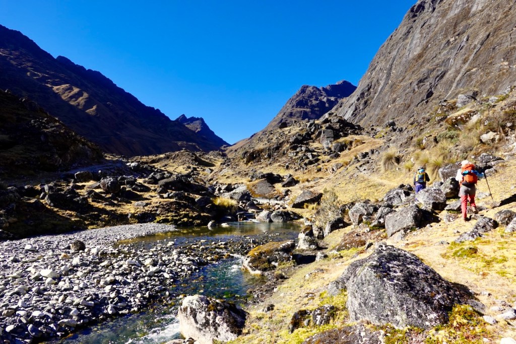 Two hikers walking along a rocky riverbank in a mountainous landscape under a clear blue sky.