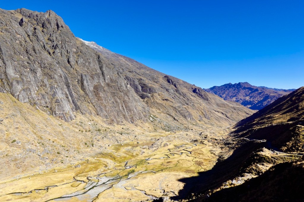 A panoramic view of a vast valley surrounded by steep mountains under a clear blue sky, featuring a winding river and dry grasslands.