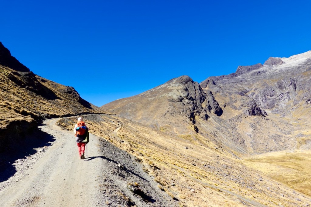 A hiker walking along a dirt trail in a mountainous landscape under a clear blue sky.