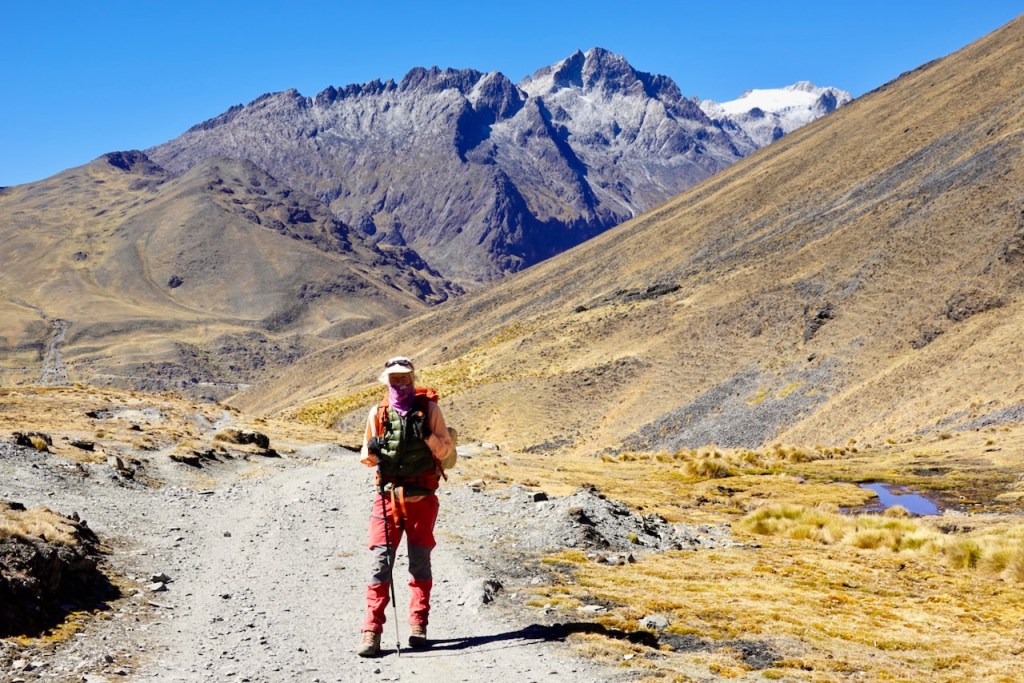 A hiker walking on a rugged mountain trail, surrounded by steep slopes and distant snow-capped peaks under a clear blue sky.