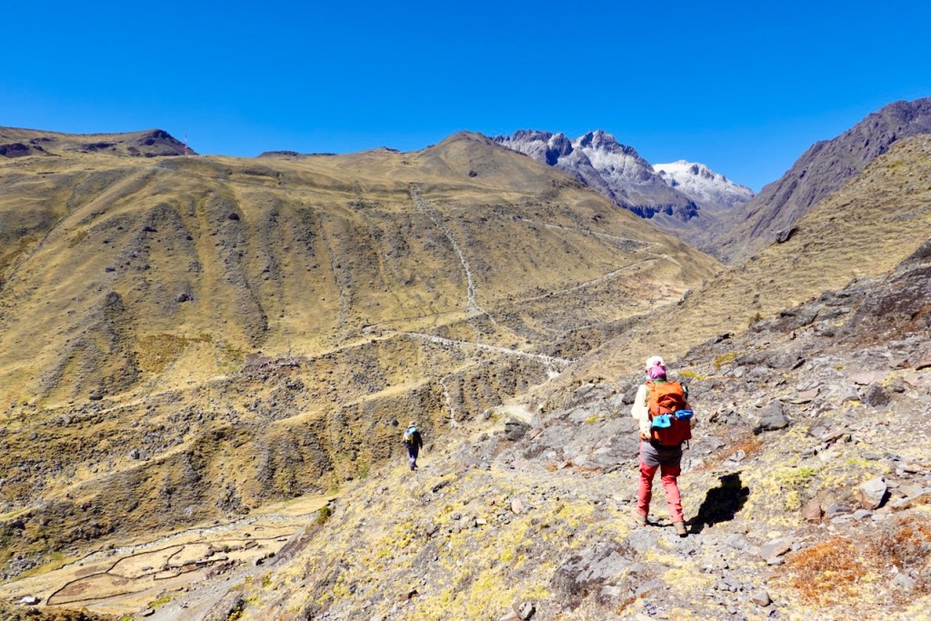 A hiker with an orange backpack standing on rocky terrain, overlooking a valley with rugged hills and snow-capped mountains in the distance under a clear blue sky.