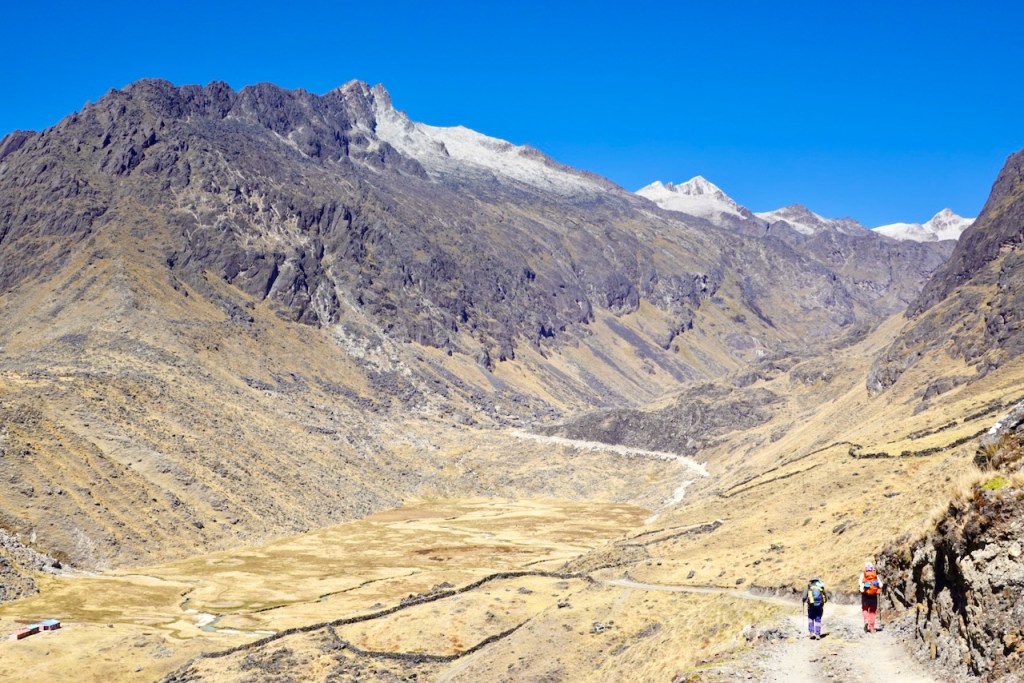 Two hikers walking along a dirt path in a mountainous landscape with steep slopes and a clear blue sky.