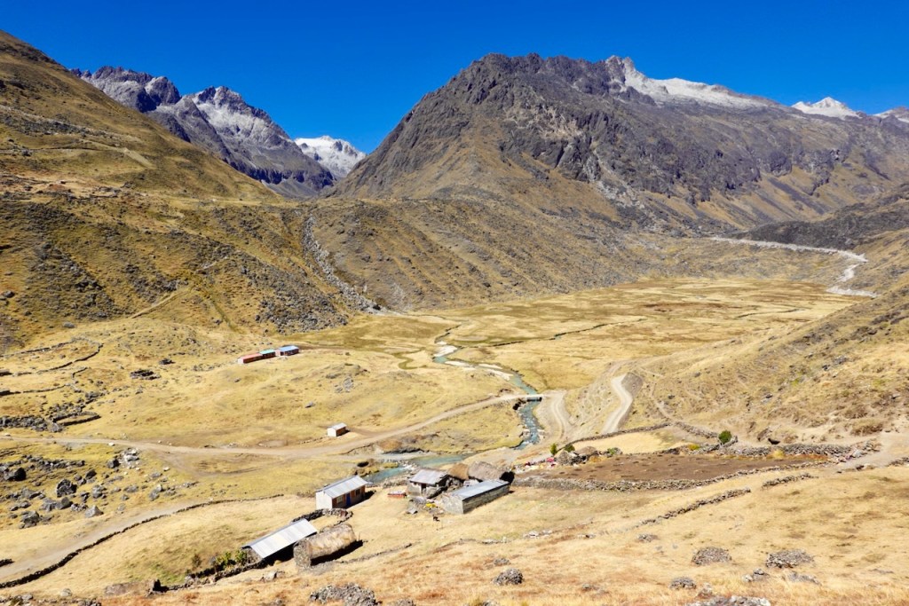 A panoramic view of a mountainous landscape featuring dry, grassy terrain, a winding stream, and scattered buildings in a remote area.