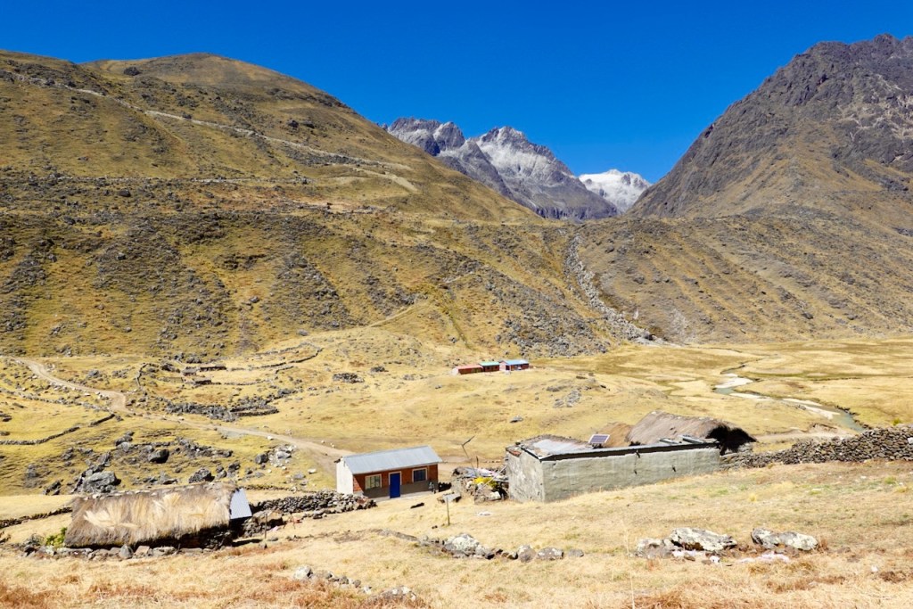 Scenic mountain landscape featuring rocky hills, grassy plains, and structures including a thatched-roof house and a simple building in a rural setting.