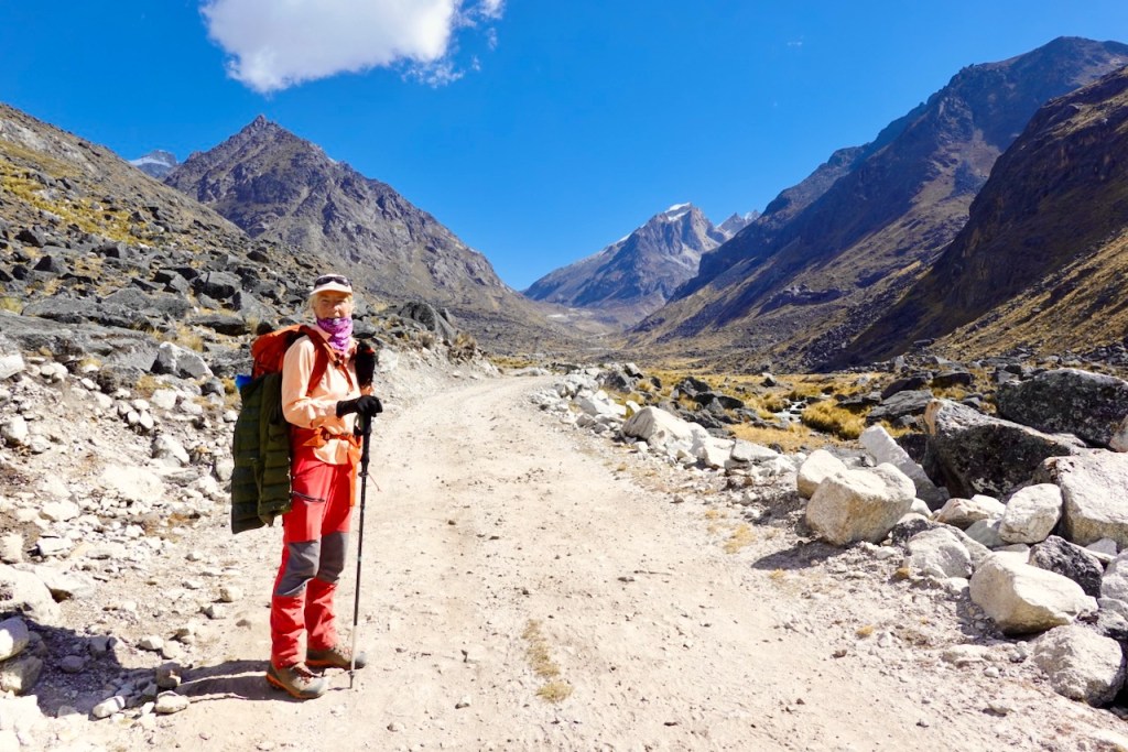 Hiker standing on a dirt path surrounded by mountains and rocky terrain, wearing outdoor gear and holding a trekking pole.