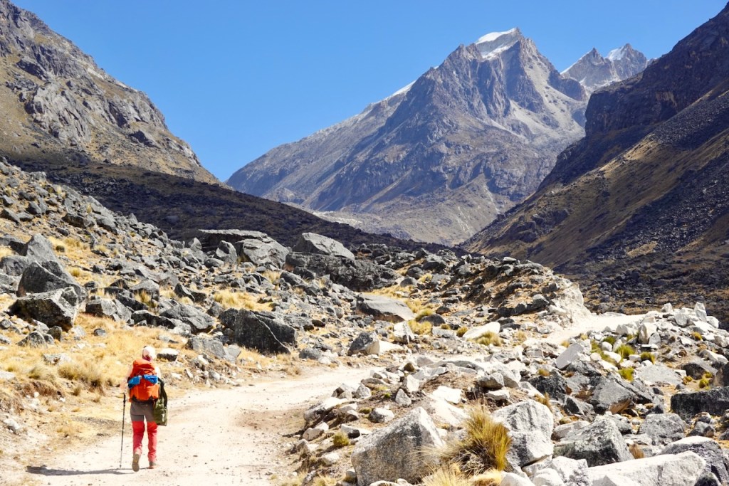 A hiker in red pants and an orange jacket walking along a rocky path in a mountainous landscape with blue skies.