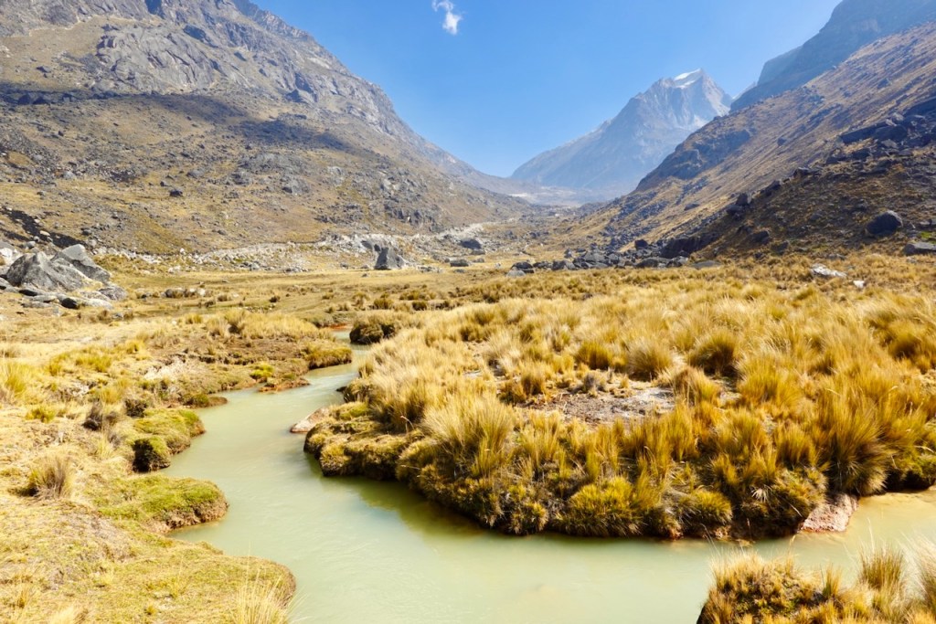 Scenic view of a grassy valley with a winding stream surrounded by rocky mountains under a clear blue sky.