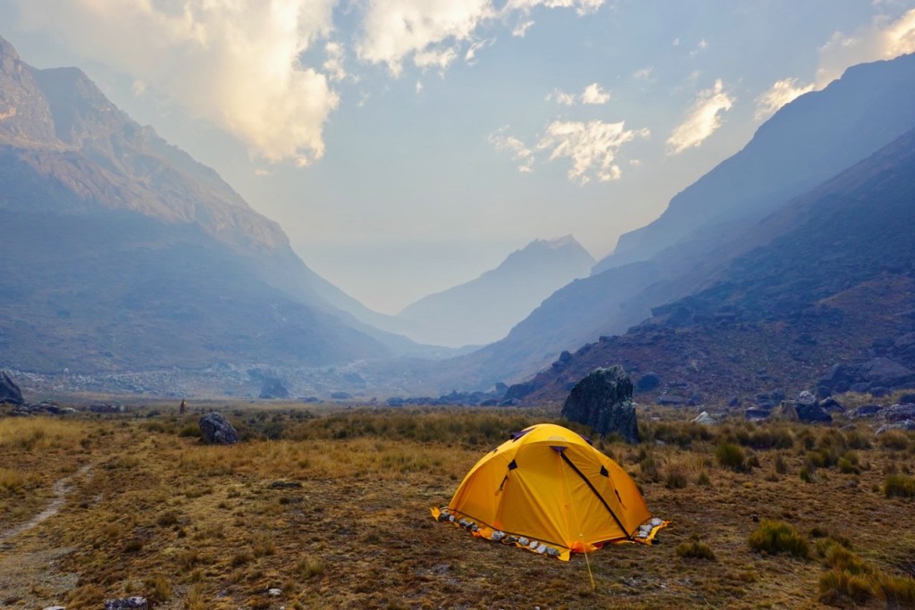 A yellow tent set up in a rugged valley surrounded by steep mountains under a cloudy sky.