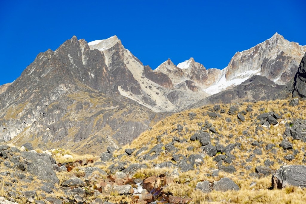 A panoramic view of rugged mountain peaks under a clear blue sky, showcasing rocky terrain and patches of snow on the summit.