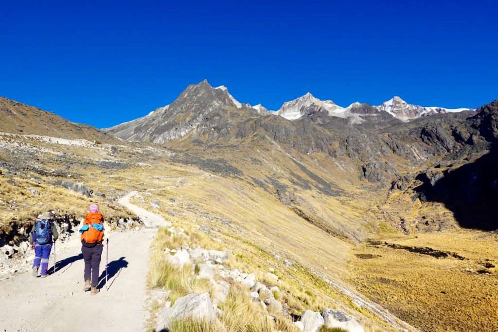 Two hikers walking on a mountain trail in a mountainous landscape under a clear blue sky.