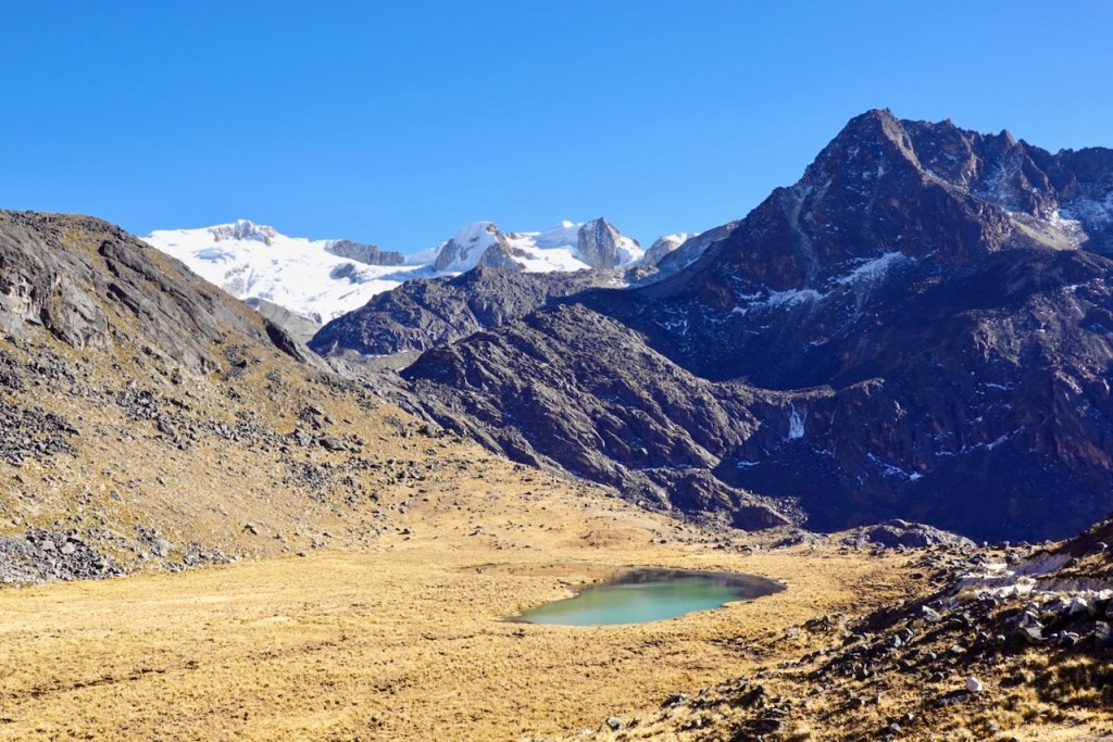 A scenic view of a mountainous landscape featuring snow-capped peaks, a rocky terrain, and a clear turquoise lake surrounded by golden grass.