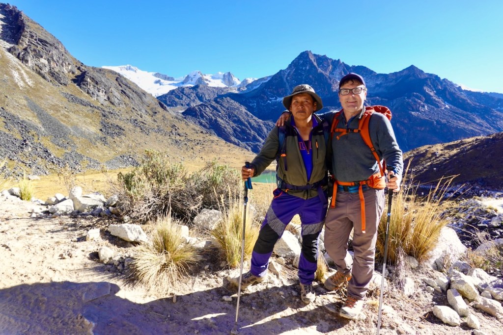 Two hikers pose for a photo on a mountain trail, with towering peaks and a clear blue sky in the background.
