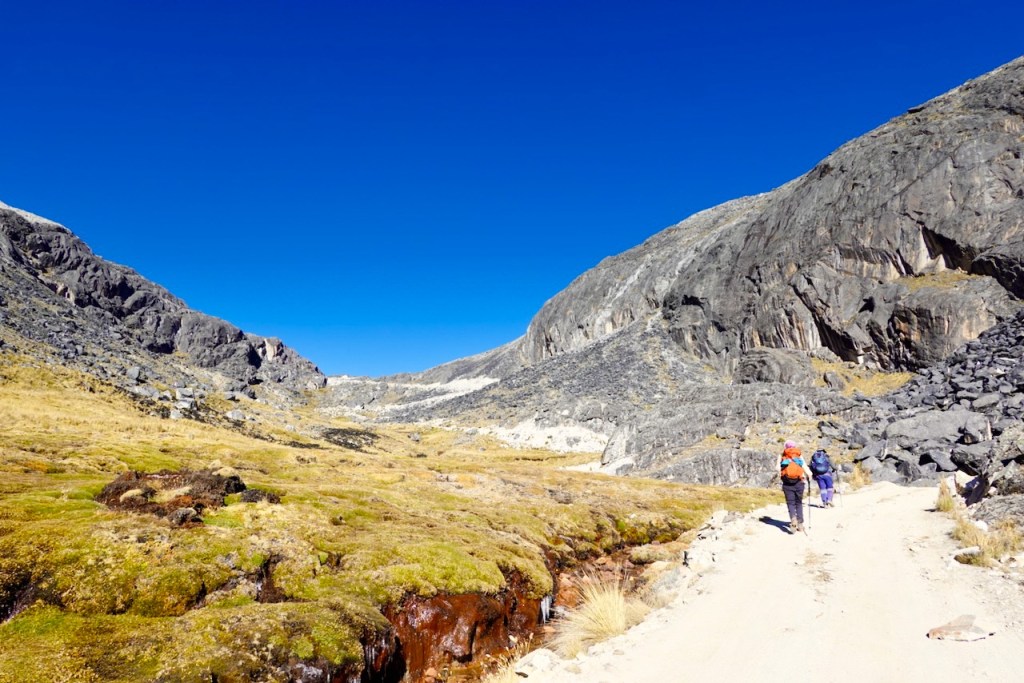 Two hikers walking on a dirt path through a rocky landscape with mountains and a clear blue sky.