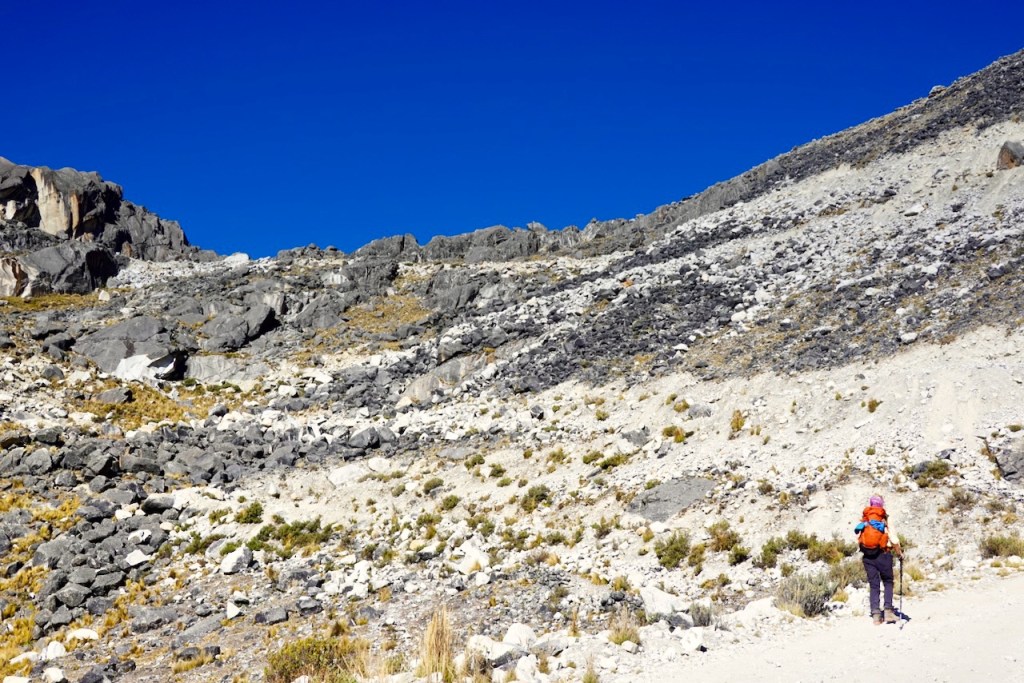 A hiker walking on a rocky trail surrounded by steep mountains and a clear blue sky.