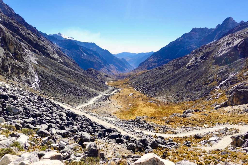 A panoramic view of a mountain valley with rocky terrain and a winding dirt road, surrounded by steep mountains under a clear blue sky.