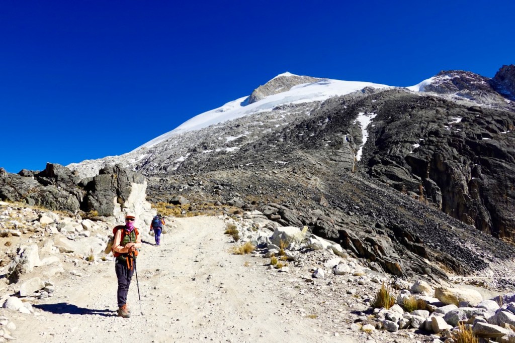 Two hikers walking on a rocky trail with snow-capped mountains and a clear blue sky in the background.