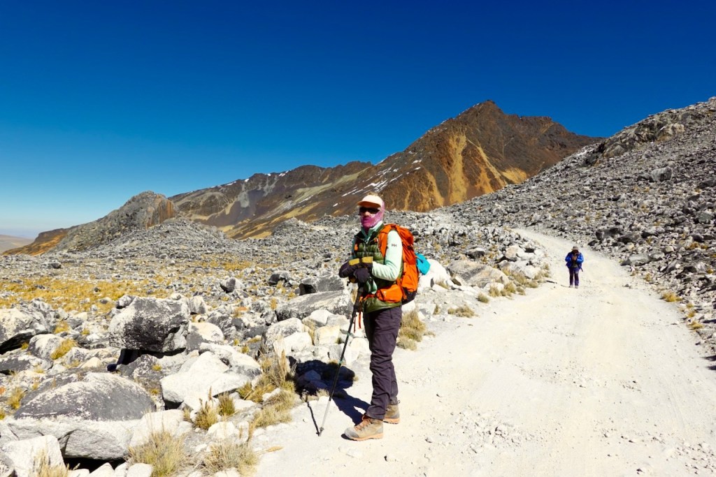 A hiker stands on a rocky path in a mountainous area, wearing a backpack and trekking poles, with a fellow hiker visible in the background.