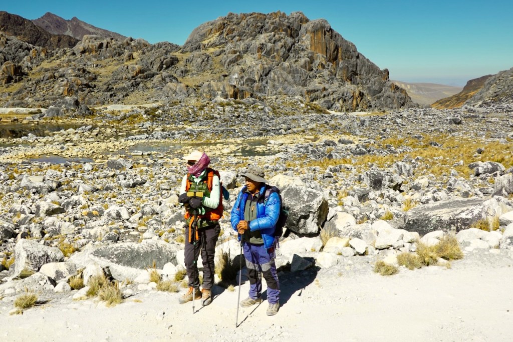 Two hikers standing on a rocky mountain trail with steep cliffs and distant peaks in the background.