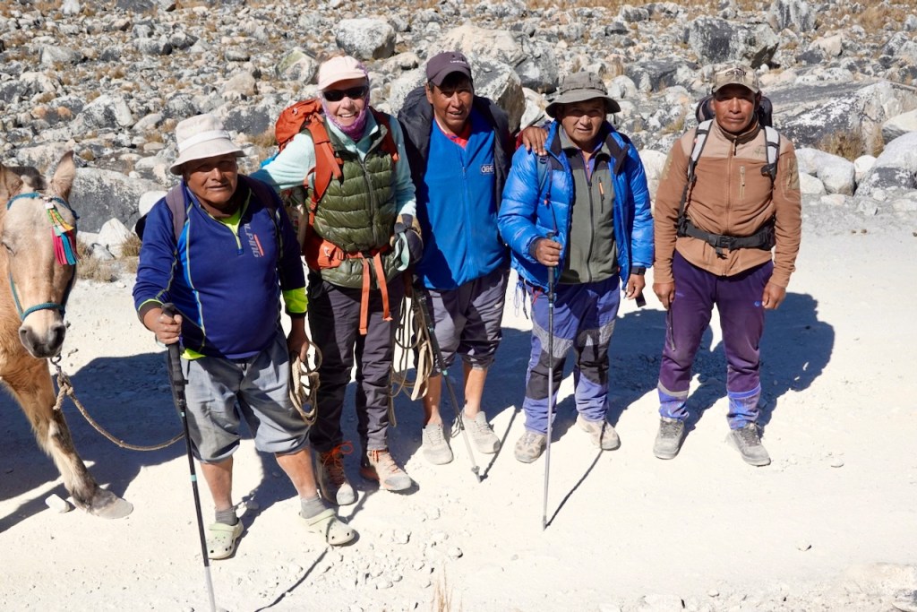 A group of five men standing together on a rocky outdoor trail, two holding trekking poles, with a horse on the left side.
