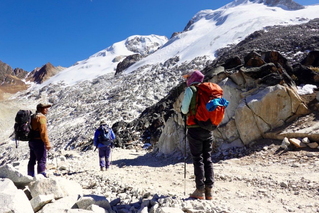 Three hikers trekking along a rocky trail in a mountainous region with snow-capped peaks in the background.