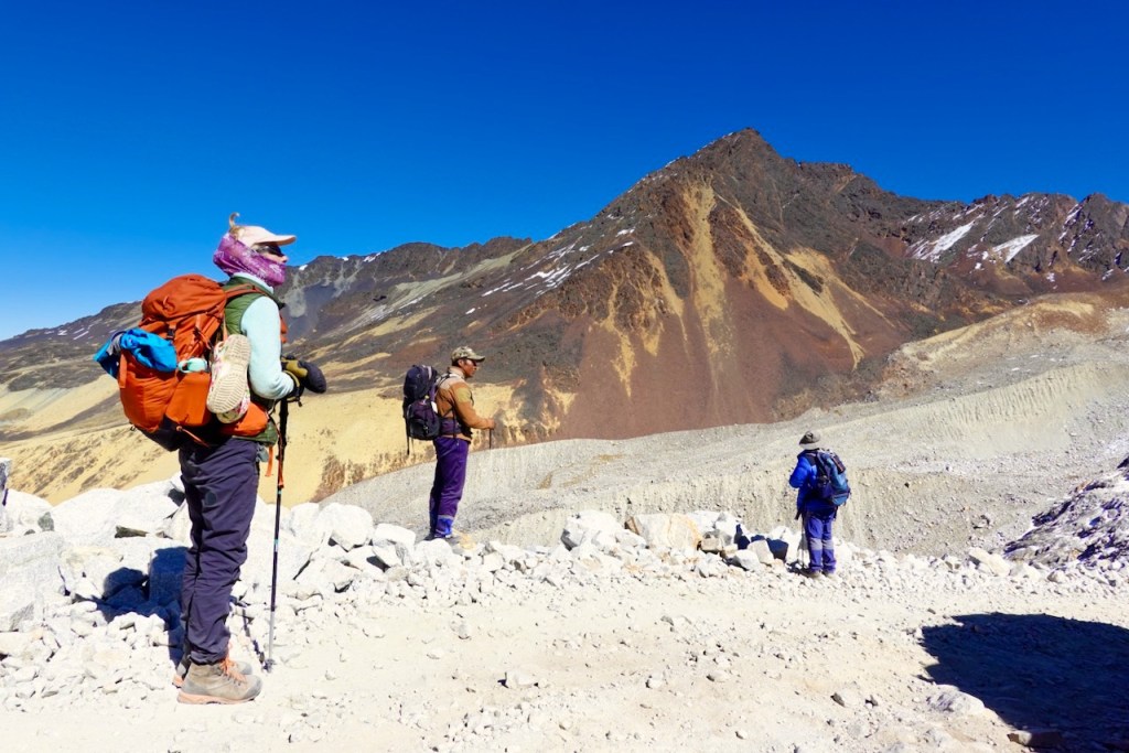 Three hikers on a rocky mountain trail, with a prominent mountain peak in the background under a clear blue sky.