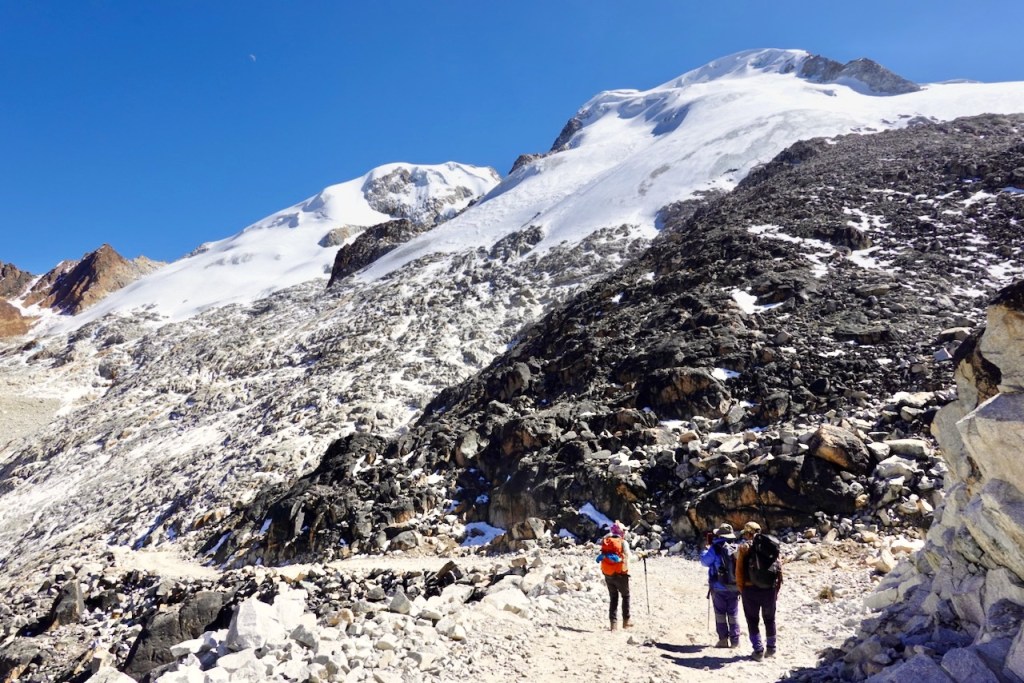 Two hikers walking on a rocky trail with snow-covered mountains in the background under a clear blue sky.