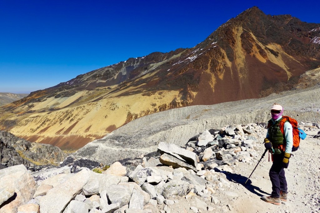 A hiker standing on a rocky mountain trail with a colorful backpack, surrounded by vibrant, multi-colored slopes under a clear blue sky.
