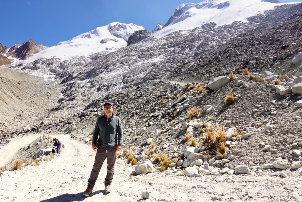 A person standing on a rocky mountain path with snow-capped peaks in the background.