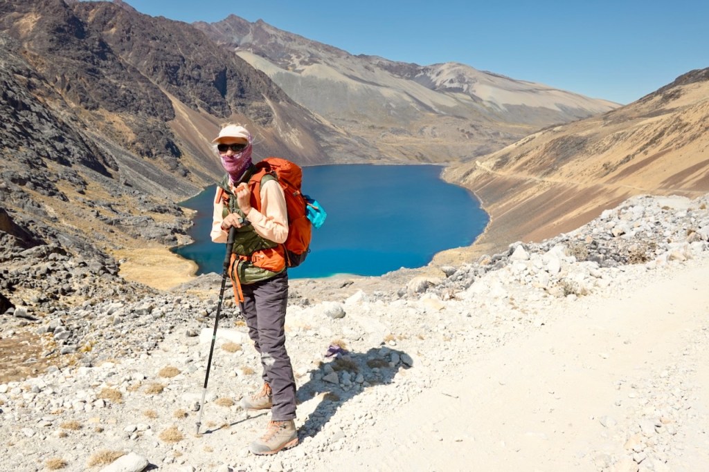 A hiker standing on a rocky path overlooking a bright blue lake surrounded by mountainous terrain.