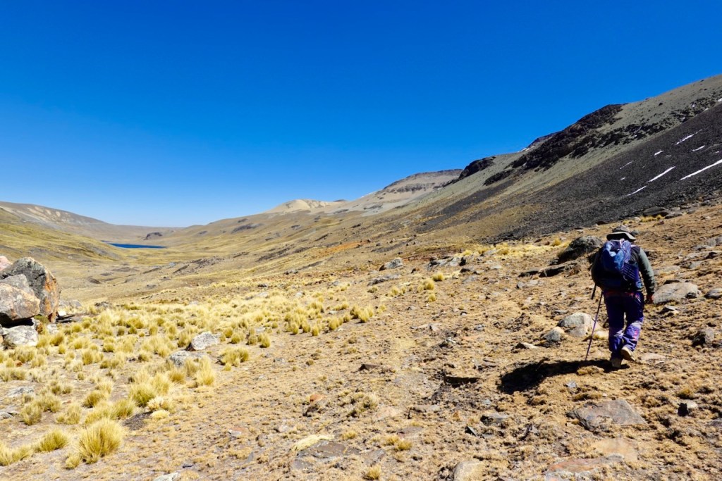 A hiker walking through a mountainous landscape with a clear blue sky and a distant lake visible in the valley.