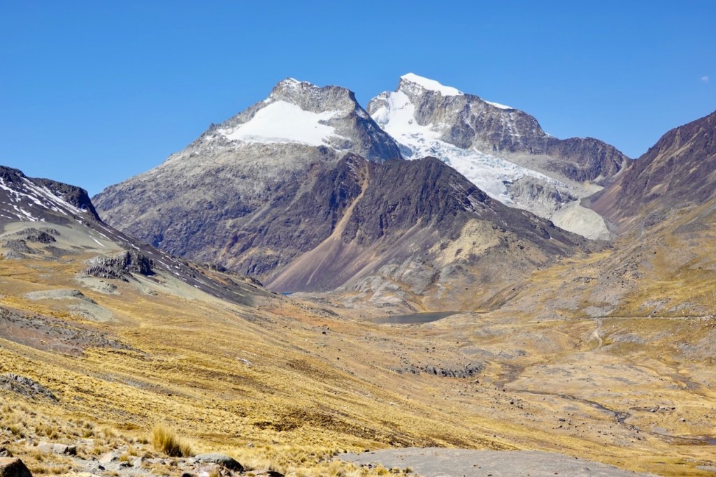 A panoramic view of mountains with snow-capped peaks against a clear blue sky, surrounded by rocky terrain and dry grass.