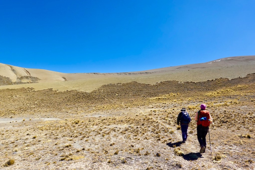 Two hikers walking on a rocky trail in a mountainous landscape under a clear blue sky.