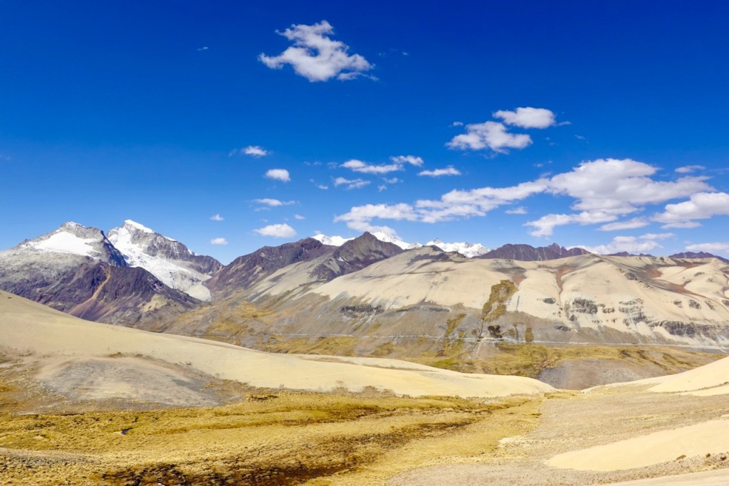 Panoramic view of mountainous landscape with snow-capped peaks and blue sky dotted with clouds, featuring rugged terrain and rock formations.