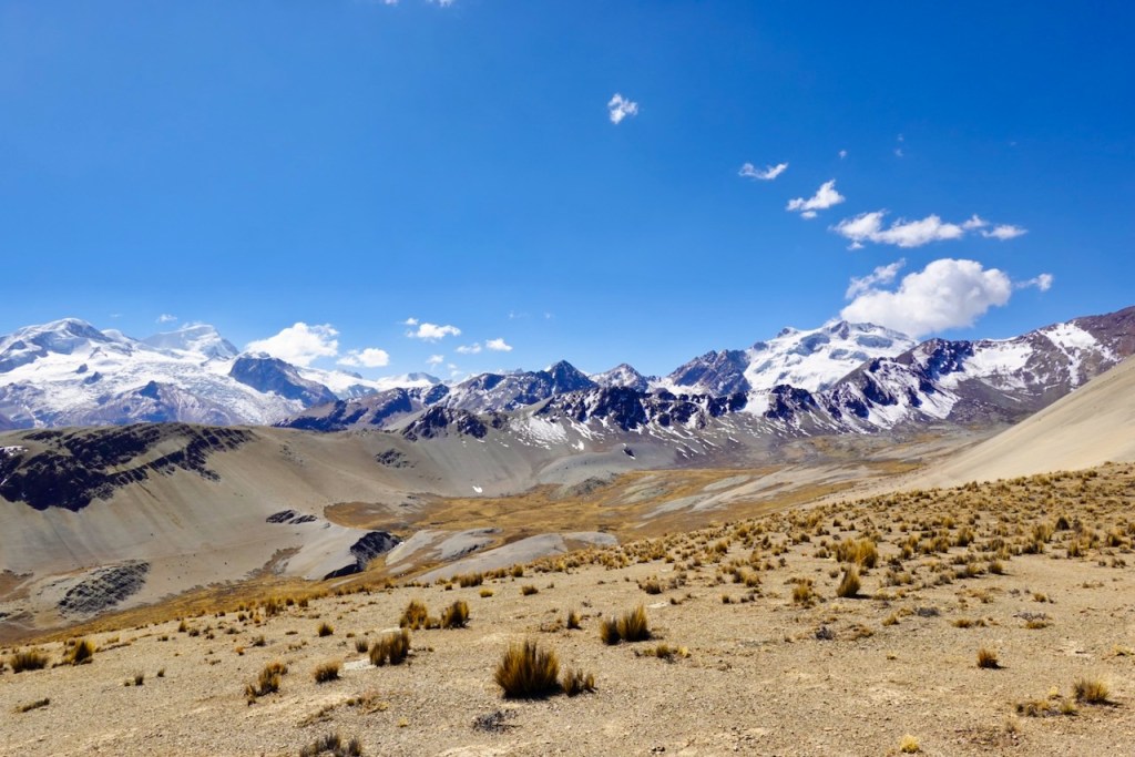 A panoramic view of a mountainous landscape with snow-capped peaks under a clear blue sky. The foreground features arid terrain and sparse vegetation.