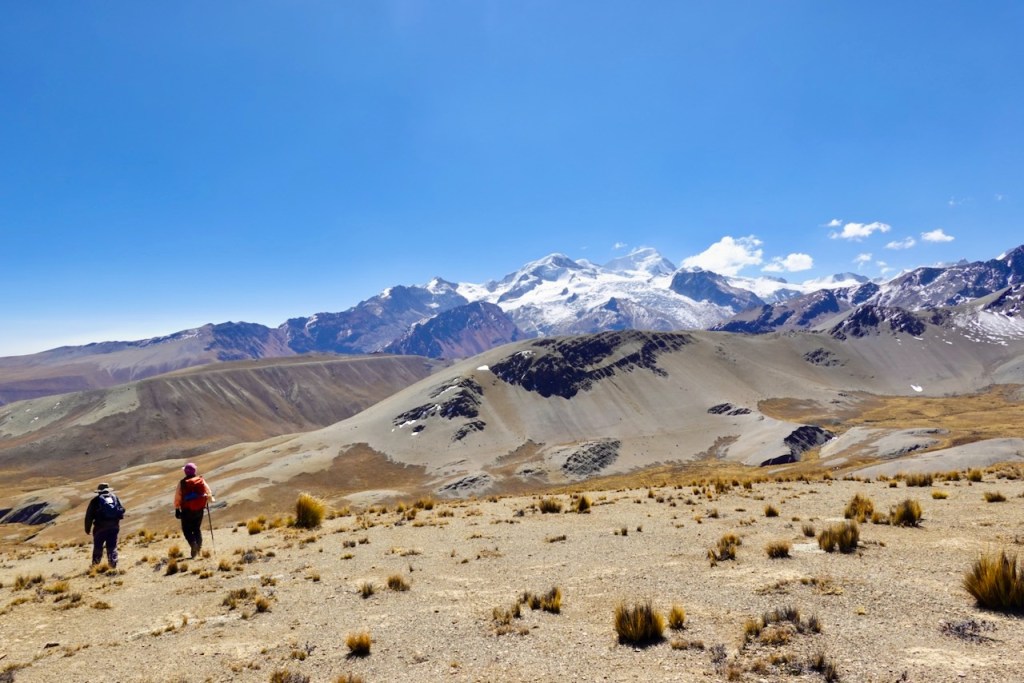 Two hikers walking on a rocky terrain with mountains in the background under a clear blue sky.