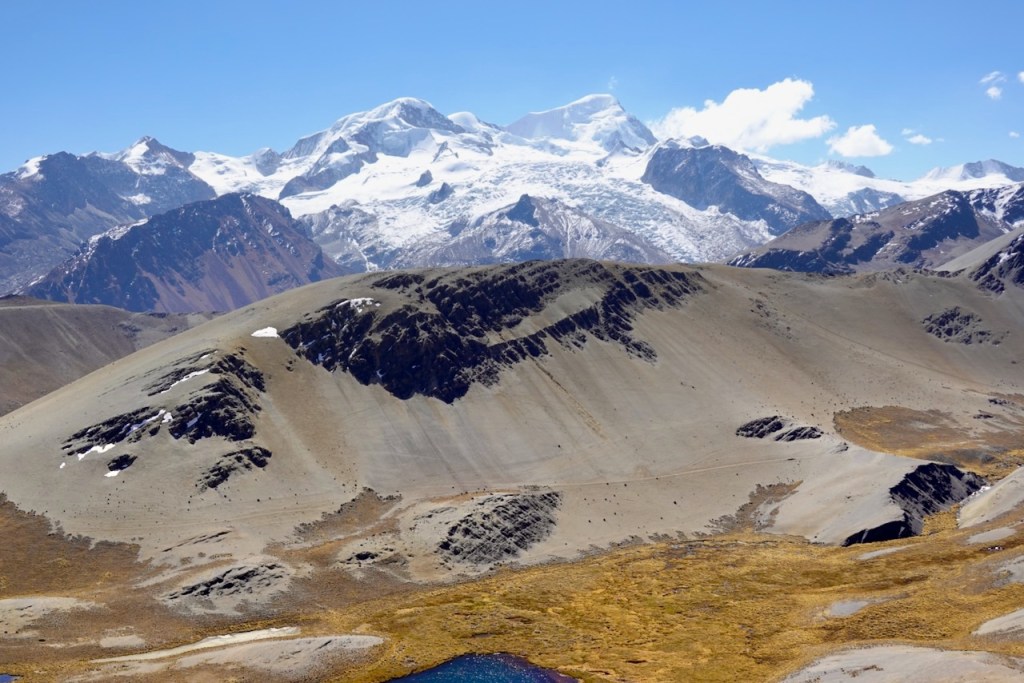 A panoramic view of snow-capped mountains under a clear blue sky, with a rugged landscape featuring brown hills and patches of greenery in the foreground.
