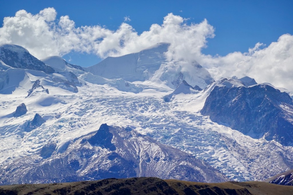 A breathtaking view of snowy mountain peaks under a blue sky with fluffy clouds, showcasing glaciers and rocky terrain.