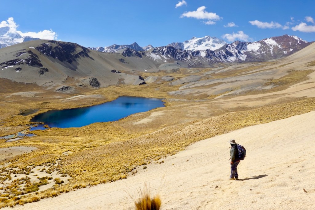 A hiker standing on a sandy slope overlooking a vibrant blue lake surrounded by mountains under a clear sky.