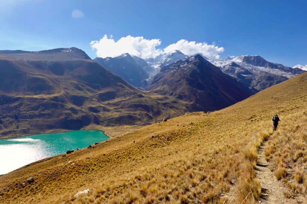 A hiker walking along a narrow trail in a mountainous landscape, with greenish-blue lake visible below and snow-capped peaks in the background under a clear blue sky.