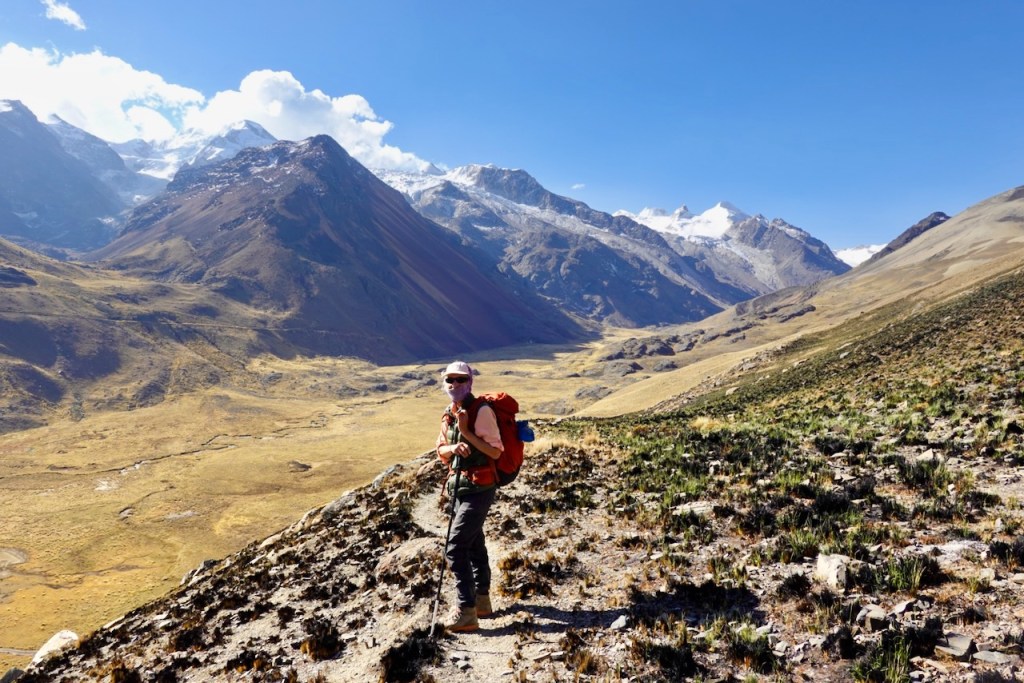 A hiker with a backpack stands on a rocky path overlooking a vast mountainous landscape under a blue sky.