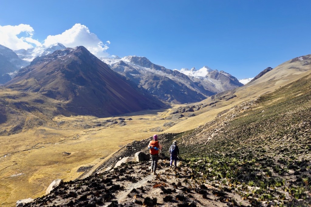Two hikers walking along a rocky trail in a mountainous landscape with grass and sparse vegetation, under a clear blue sky.